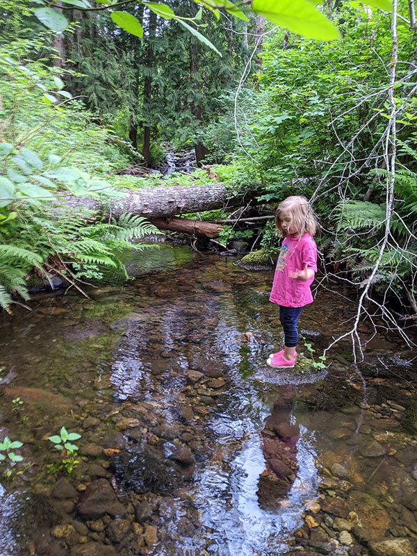 A reflection in the water of a four year old child dressed in pink water shoes.